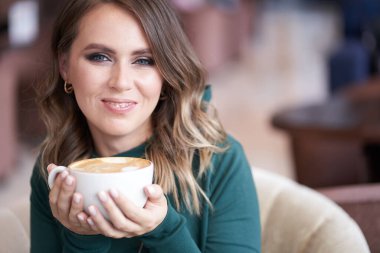 Güzel bir saç stili ile mutlu güzel bir kız, elinde bir fincan kahve, yeşil balıkçı yaka, bir kafede oturuyor, kameraya bakıyor ve gülümsüyor.