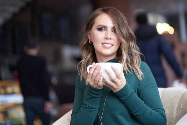Saç stili ve elinde bir fincan kahve olan güzel bir kadın bir kafede oturuyor. Doğrudan kameraya bakıp gülümsüyor. — Stok fotoğraf