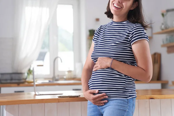 Eine schwangere Frau wickelt ihre Arme um ihren Bauch. Eine Frau im dritten Schwangerschaftsdrittel steht in der Küche. Das Konzept des Wartens auf ein Kind. — Stockfoto