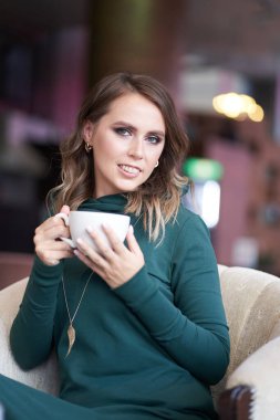 Bir fincan kahve tutan, saç stili olan güzel bir kadın, bir kafede oturuyor. Gülümseyerek doğrudan kameraya bakıyor.