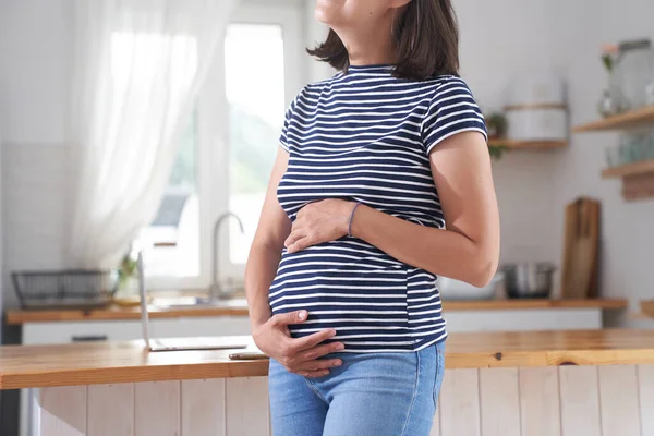 Eine schwangere Frau wickelt ihre Arme um ihren Bauch. Eine Frau im dritten Schwangerschaftsdrittel steht in der Küche. Das Konzept des Wartens auf ein Kind. — Stockfoto