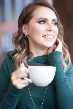 İyimser, güzel bir esmer kadının fotoğrafı. Saç stili, elinde fincan, yeşil balıkçı yaka bir kazak, kafede oturuyor.