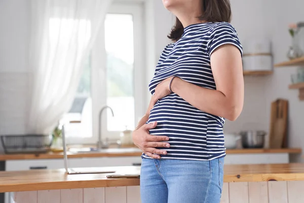 Eine junge schwangere Frau hält ihren Bauch mit den Händen. Eine brünette Frau im dritten Schwangerschaftsdrittel steht in der Küche und umarmt ihren Bauch. Das Konzept des Wartens auf ein Kind. — Stockfoto
