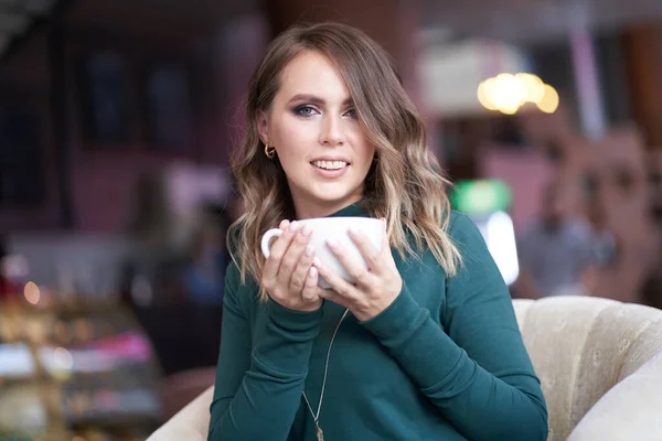 Saç stili ve elinde bir fincan kahve olan güzel bir kadın bir kafede oturuyor. Gülümseyerek doğrudan kameraya bakıyor. — Stok fotoğraf
