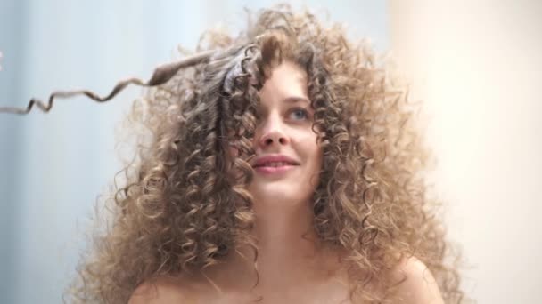 Close-up af en smuk kvinde med krøllet hår, hendes hår bliver gjort, hun blæser på en streng af hår ser på kameraet – Stock-video