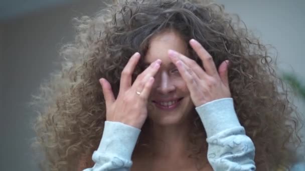 Slow motion. Close-up of a beautiful young woman with curly hair, she looks at the camera smiling, touches her hair with her hands — Stockvideo