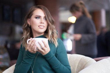 Saç stili ve elinde bir fincan kahve olan güzel bir kadın bir kafede oturuyor. Gülümseyerek yüzünü çeviriyor.