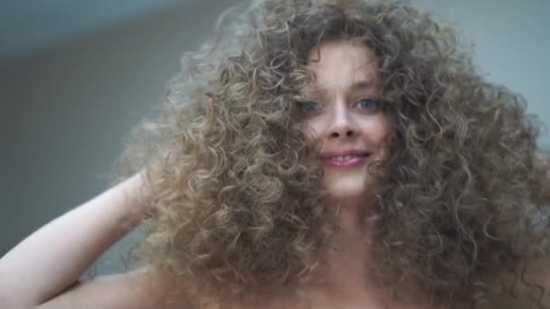 Slow motion. Close-up of a beautiful young woman with curly hair, she looks at the camera smiling, touches her hair with her hands — Stockvideo