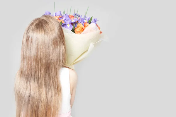 La niña sostiene un ramo de flores en sus manos. Se para con la espalda a la cámara sobre un fondo gris claro —  Fotos de Stock