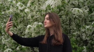 Gülümseyen genç bir kız parkta, çiçek açan beyaz bir elma ağacının arka planında kendi fotoğraflarını çekiyor. Gençler rüzgar sırasında akıllı telefondan selfie çeker.
