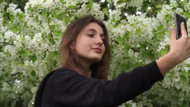 Крупный план молодой девушки, фотографирующей себя на фоне цветущей белой яблони. Подросток делает селфи на смартфоне. — стоковое видео
