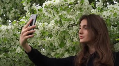 Gülümseyen genç bir kız parkta, çiçek açan beyaz bir elma ağacının arka planında kendi fotoğraflarını çekiyor. Gençler rüzgar sırasında akıllı telefondan selfie çeker.