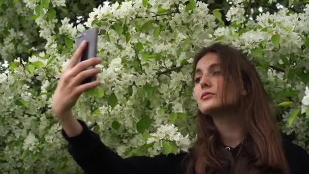 Uśmiechnięta młoda dziewczyna robi sobie zdjęcia w parku na tle kwitnącej białej jabłoni. Nastolatek robi sobie selfie na smartfonie. — Wideo stockowe