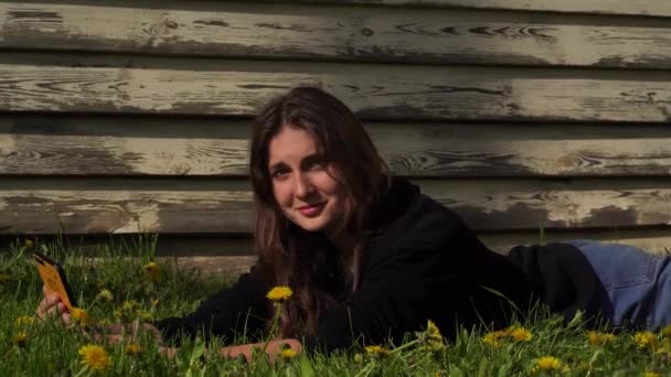 Egy tinédzser fényképeket készít magáról egy okostelefonon.Egy mosolygó fiatal lány fekszik a zöld füvön a parkban.. — Stock videók