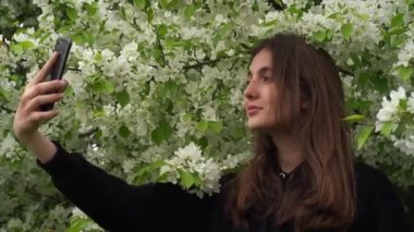Bir genç rüzgar sırasında akıllı telefondan selfie çeker. Gülümseyen genç bir kız parkta, çiçek açan beyaz bir elma ağacının arka planında kendi fotoğraflarını çekiyor..