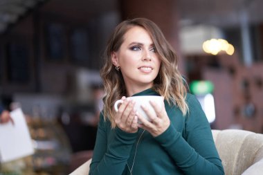 Saç stili ve elinde bir fincan kahve olan güzel bir kadın bir kafede oturuyor. Gülümseyerek yüzünü çeviriyor.