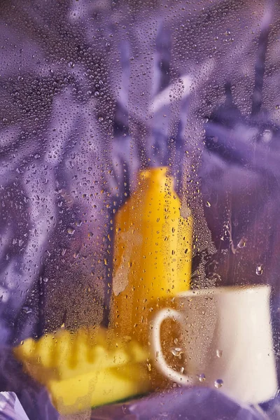 Household cleaning products and kitchen accessories. A plastic cup and a bottle with a sponge on a lilac background. View through wet glass.
