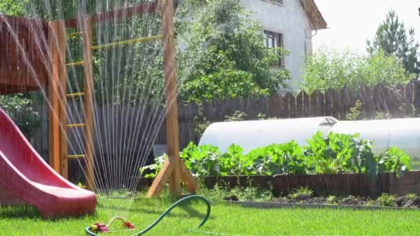 Gyerekjátszótér a hátsó kertben. A locsoló egy nyári napon öntözi a gyepet a csúszda közelében. — Stock videók