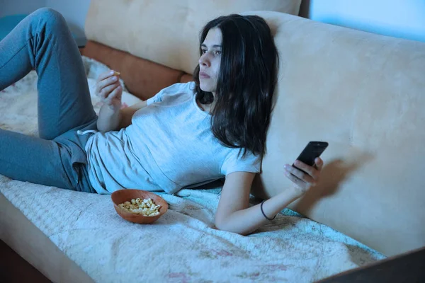 Un'immagine ravvicinata di una giovane donna triste che mangia popcorn. Utilizza il telecomando dalla TV a casa sul divano Immagini Stock Royalty Free