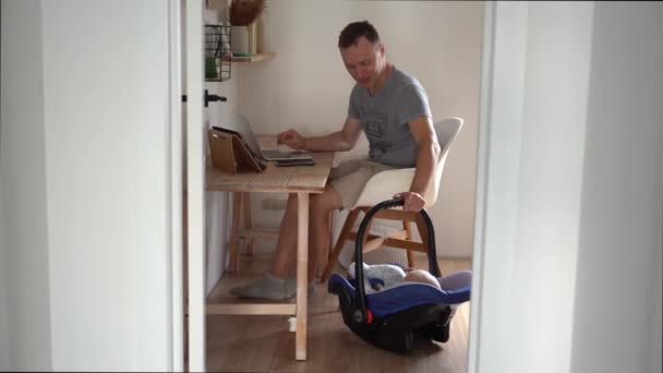 ハンサムなフリーランスの男が現代のラップトップで働いています。子供の父親は家で働いて息子をクレードルに詰め込む. — ストック動画