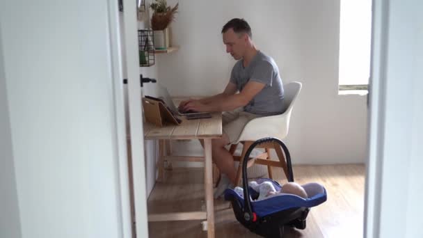 ハンサムな若い男が現代のノートパソコンで働いている。幼い父親がクレードルで息子の隣の家で働いている — ストック動画