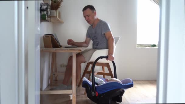 ハンサムなフリーランスの男が現代のラップトップで働いています。子供の父親は家で働いて息子をクレードルに詰め込む. — ストック動画