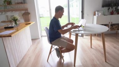 Genç bir erkek serbest çalışan akıllı telefondan bir şeye bakıyor ve dizüstü bilgisayarda yazıyor. Modern bir dizüstü bilgisayarla evde otururken uzaktan çalışıyor..