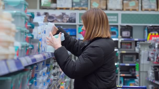 Γυναίκα με μάσκα κορωναϊού επιλέγει αγαθά σε ένα υπερμάρκετ, το τμήμα επιτραπέζιων σκευών. Επιλεκτική εστίαση — Αρχείο Βίντεο