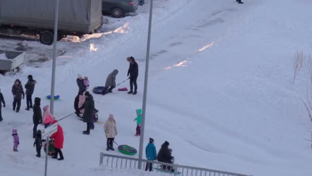 Tyumen, Rusland-23 januari 2022: Winterdag vrij. Ouders met kinderen rijden op stappen van een glijbaan in de buurt van de oever van een bevroren meer — Stockvideo