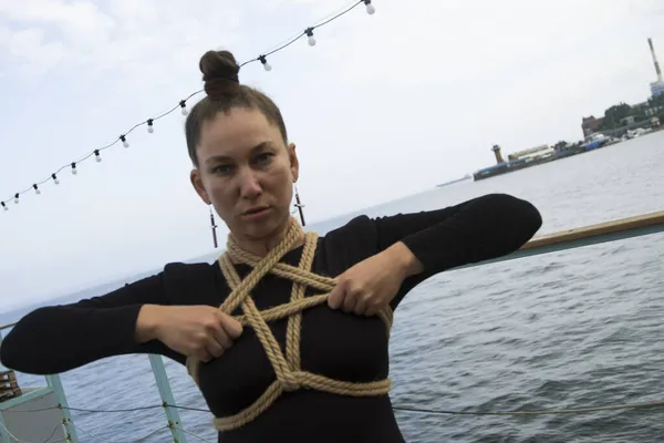Siyah dar bluzlu genç bir kız, göbeği pentagram halatıyla bağlanmış, açık güneşli bir günde deniz kenarında poz veriyor, iki kolu da göğsüne yakın, ipleri tutuyor.