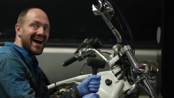 一个穿着蓝色工作服 留着短胡子 发型的机械师就在摩托车旁边 这个人一边说着摩托车的构造 一边用手做着手势 说着怎样修理它 — 图库视频影像