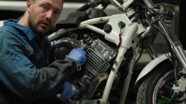 Mavi tulumlu, kısa sakallı ve saç tıraşlı bir tamirci motosikletin yanında. Adam motosikletin cihazından ve elleriyle nasıl tamir edileceğinden bahsediyor..