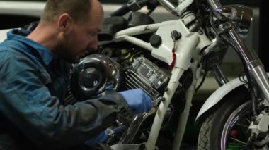 Mavi tulumlu, kısa sakallı ve saç tıraşlı bir tamirci motosikletin yanında. Adam motosikletin cihazından ve elleriyle nasıl tamir edileceğinden bahsediyor..