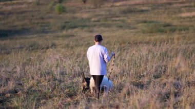 Ceketli ve başında kep olan bir genç kız tarlada üç köpekle yürüyor, köpeklerden ikisi tasmalı tutuluyor. Gün batımından önce, gökyüzü turuncu güneş ışığıyla doludur, ağır çekim.