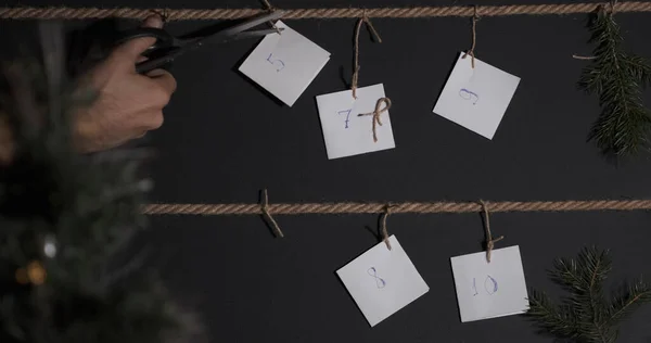 Ev yapımı DIY takvimi, siyah arka planda yaratıldı ve bir el işi teli ile bağlandı, ön planda çelenkli bir Noel ağacı var. Makas etiketlerden birini kesmek için uzanır