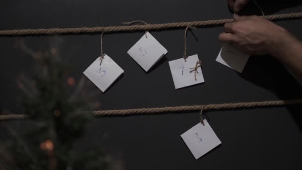 自家製Diyアドベンチャーカレンダー 黒の背景に作成され クラフト文字列でタイ 前景にはガーランドとクリスマスツリーです 男性の手は別のタグを結びます — ストック動画