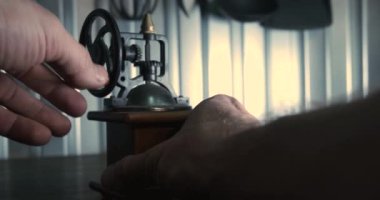 Duvarın önündeki masanın üzerinde duran küçük, mekanik, eski bir kahve öğütücüsü, erkeklerin elleri ona uzanıyor, mekanizmanın sapını çeviriyor ve geri çekiliyor.