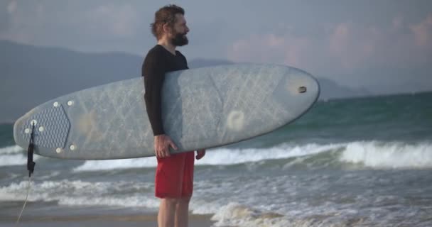 O yaştaki yakışıklı bir sporcunun portresi elinde sörf tahtasıyla sahilde duruyor ve dalgalara bakıyor. Sörften emekli oldu.. — Stok video