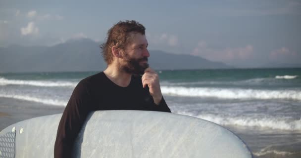 Un athlète à la retraite regarde l'océan et choisit une vague pour le surf. Portrait d'un beau sportif senior en âge de porter une planche de surf sur la plage. — Video