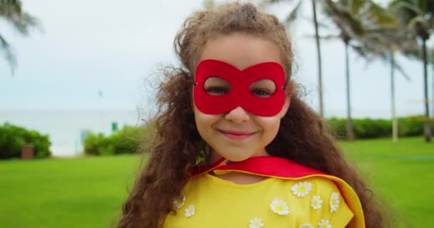 スーパーヒーローの衣装を着たかわいい面白い子供の女の子の肖像画、赤いマントと赤いマスク、公園でスーパーヒーローを演じる. — ストック動画