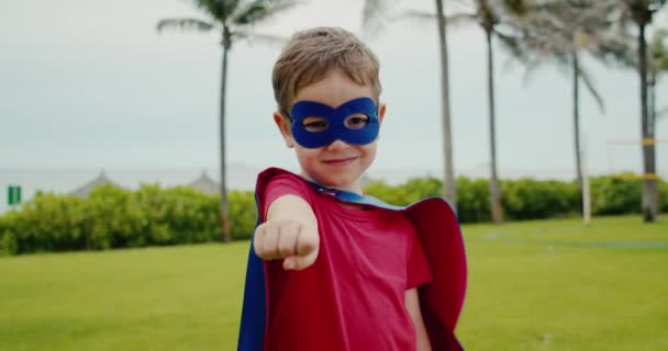 Ritratto di un simpatico ragazzo divertente in costume da supereroe, con un mantello rosso e una maschera rossa, interpreta un supereroe, dimostra la sua forza. — Video Stock