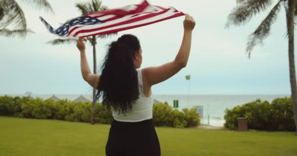 Koncepcja patriotyzmu, dnia niepodległości i wakacji - szczęśliwa uśmiechnięta młoda kobieta w T-shircie latem w parku biegnie z flagą narodową amerykańską. — Wideo stockowe