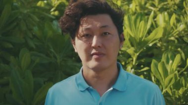 Sıradan mavi polo tişörtlü dürüst bir ifadeyle genç bir Asyalı 'nın portresi, arka plandaki tropikal ağaçlara bakıyor. Yavaş çekim 4K.