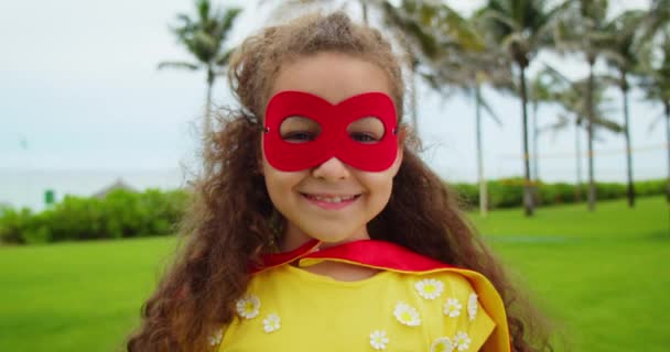 Portret słodkiej zabawnej dziewczynki w kostiumie superbohatera, w czerwonym płaszczu i czerwonej masce, grającej superbohatera w parku. — Wideo stockowe