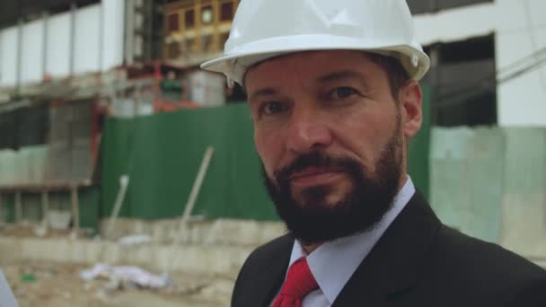 Portret starszego inżyniera z brodą, budowniczego na placu budowy, noszącego kask bezpieczeństwa, kurtkę i czerwony krawat, patrzącego w kamerę. — Wideo stockowe