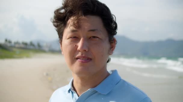 Portrait of a middle-aged asian man in a blue shirt standing on the beach by the sea against the backdrop of high mountains — Stock Video