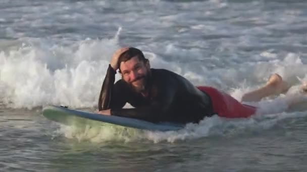 Portrait beau sportif senior avec barbe, heureux de rouler sur les vagues de la mer, couché sur la planche de surf, souriant, regardant la caméra.Athlète retraité surfe sur la mer, surf mode de vie océanique, sports extrêmes. — Video