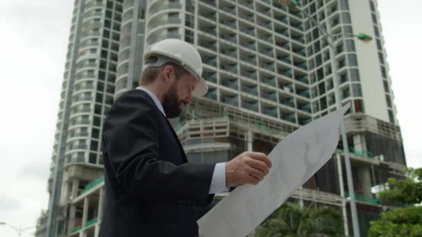 Engineer in white protective helmet checks a construction plan in new microdistrict of the city.Senior Engineer Planning Manufacture Work business suit a red jacket with construction card in his hands — Stock Video