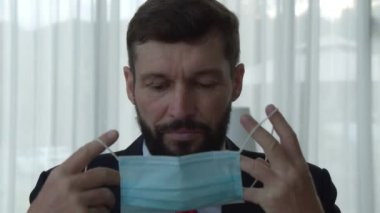 Yakışıklı, sakallı, kıdemli iş adamının portresi. Ofiste oturmuş kameraya bakıyor ve kendini korumak için tıbbi maske takıyor.