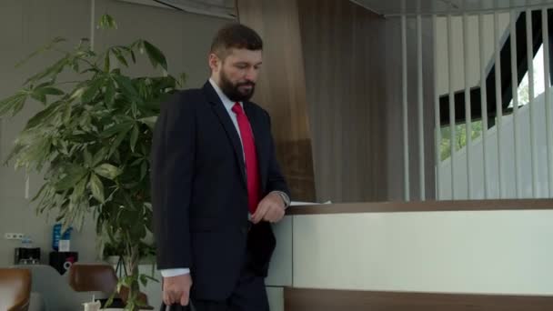 Senior zakenman met een baard in een zakenpak, een uitvoerend directeur met een koffer in zijn handen staat bij de receptie te wachten op een administrateur.Baard zakenman kijkt op zijn horloge — Stockvideo